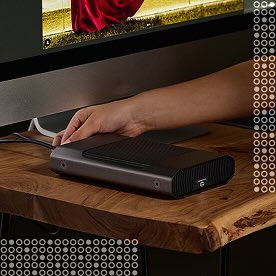 A hand plugs a cable into a black external storage device on a wooden desk, with a computer monitor in the background.