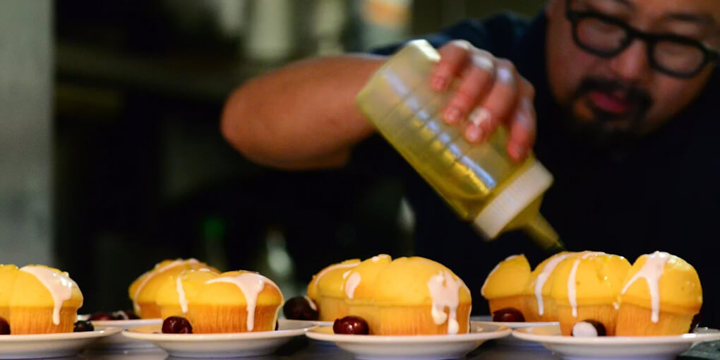A chef drizzles some oil on a grill while muffins sit in the foreground.