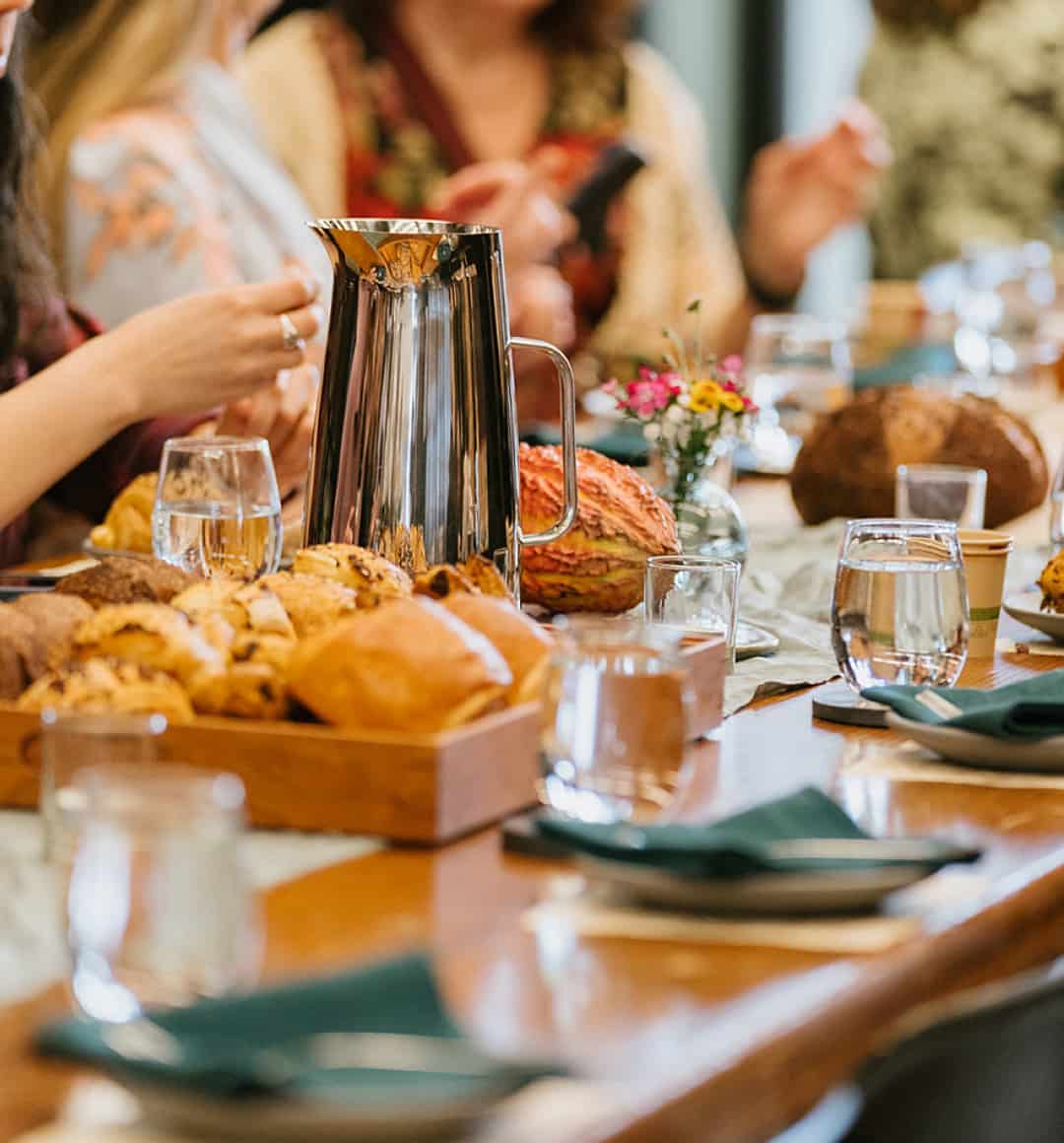A group of guests are seated around a table at the factory. On the table sit freshly baked pastries. 