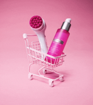 A GIF of a tiny pink shopping cart filled with a skin exfoliator tool and beauty serum.