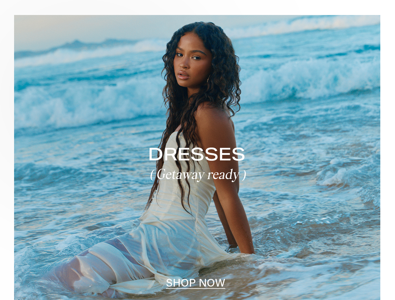 Woman with long wavy hair in a wet, flowing cream dress sits in blue ocean waves. Promoting stylish getaway dresses for your next vacation. Shop now.