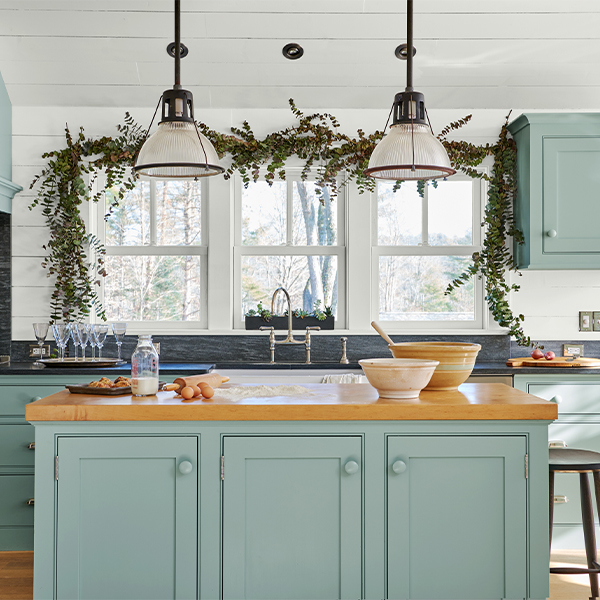 A kitchen with teal-painted cabinets, a larger butcher block island, and holiday swag draped over the windowsill.