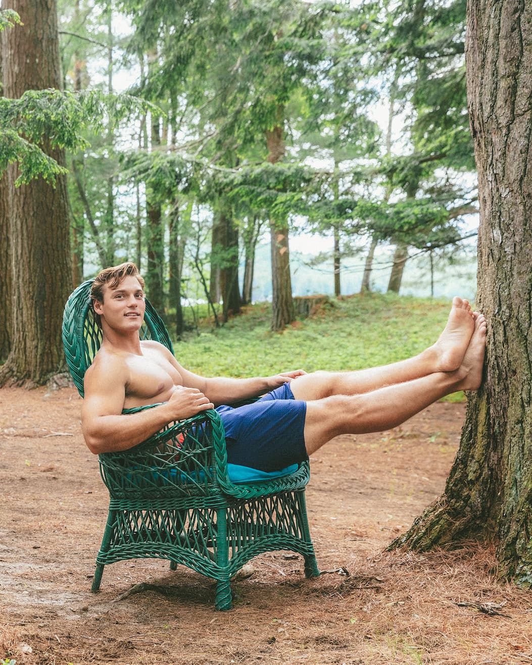 Shirtless man wearing navy blue lounge shorts sitting on a chair in the woods