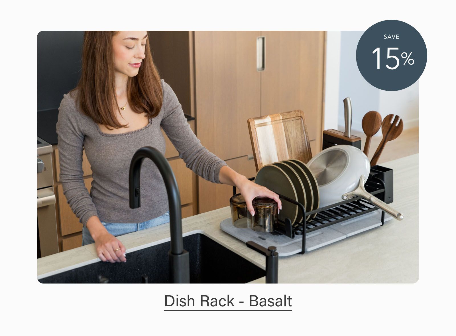 Woman placing a glass on a black Dorai Dish Rack with a gray quick-drying stone base on a modern kitchen countertop. Badge indicates 15% savings. Text reads: "Dish Rack - Basalt."