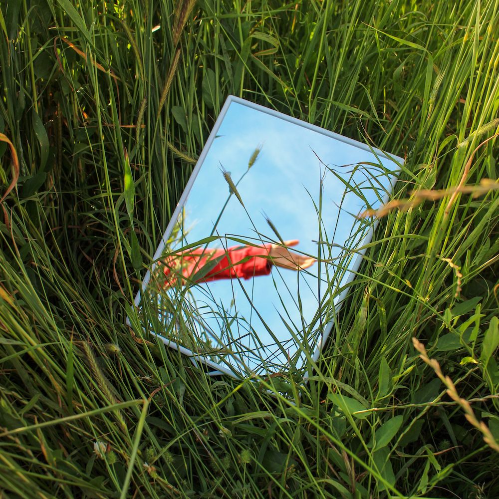 A small mirror laying in the grass reflects a person's hand in front of a blue sky. 