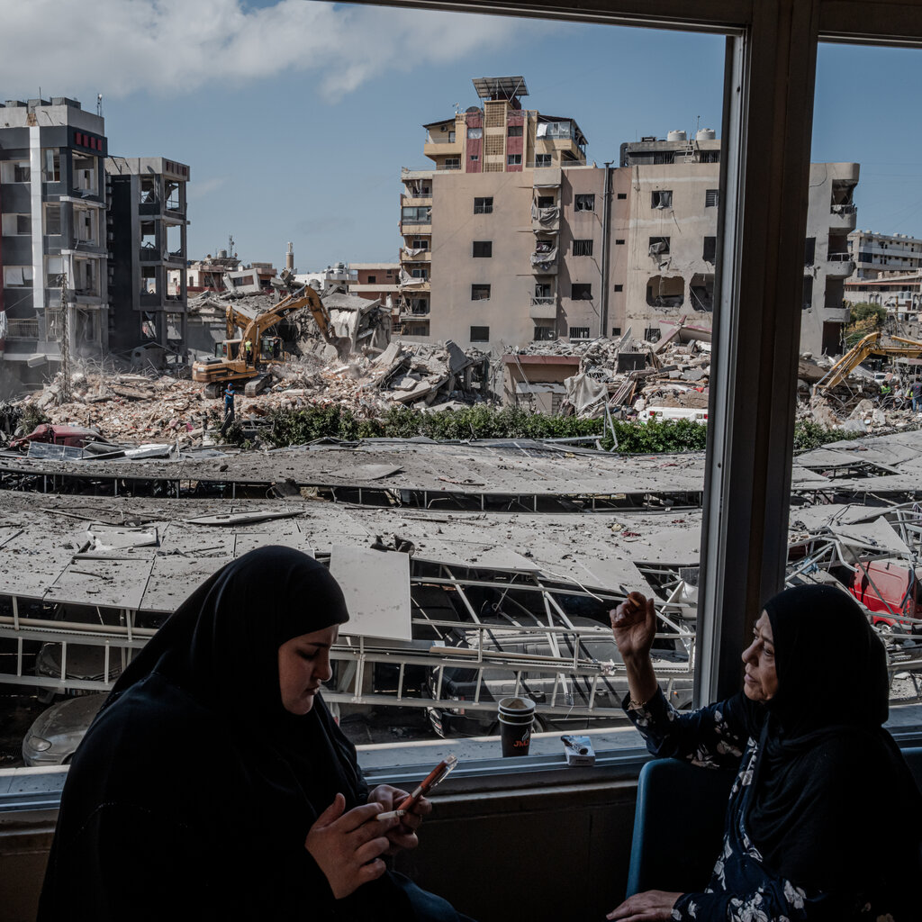Two people sit indoors looking out a window at a destroyed city. Buildings are reduced to rubble with excavators working in the debris under a blue sky.