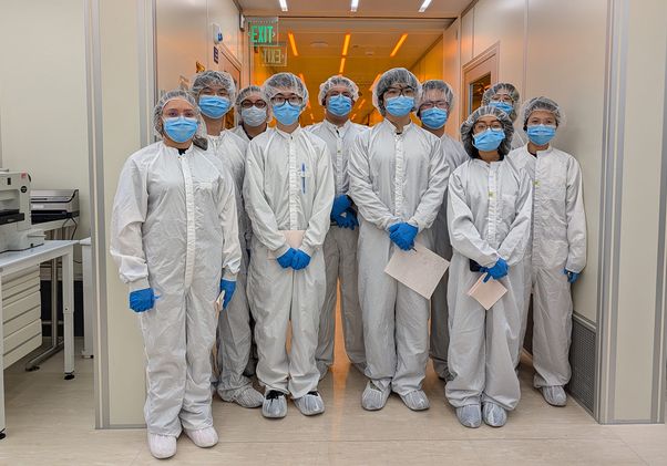 Group of 10 people in cleanroom suits, masks, gloves, and hairnets standing in a laboratory corridor, posing together.