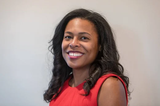 A professional headshot of a Black woman with long, wavy black hair wearing a sleeveless red dress. She is smiling, revealing straight white teeth, and looking directly at the camera. The background is a neutral light gray, softly blurred.