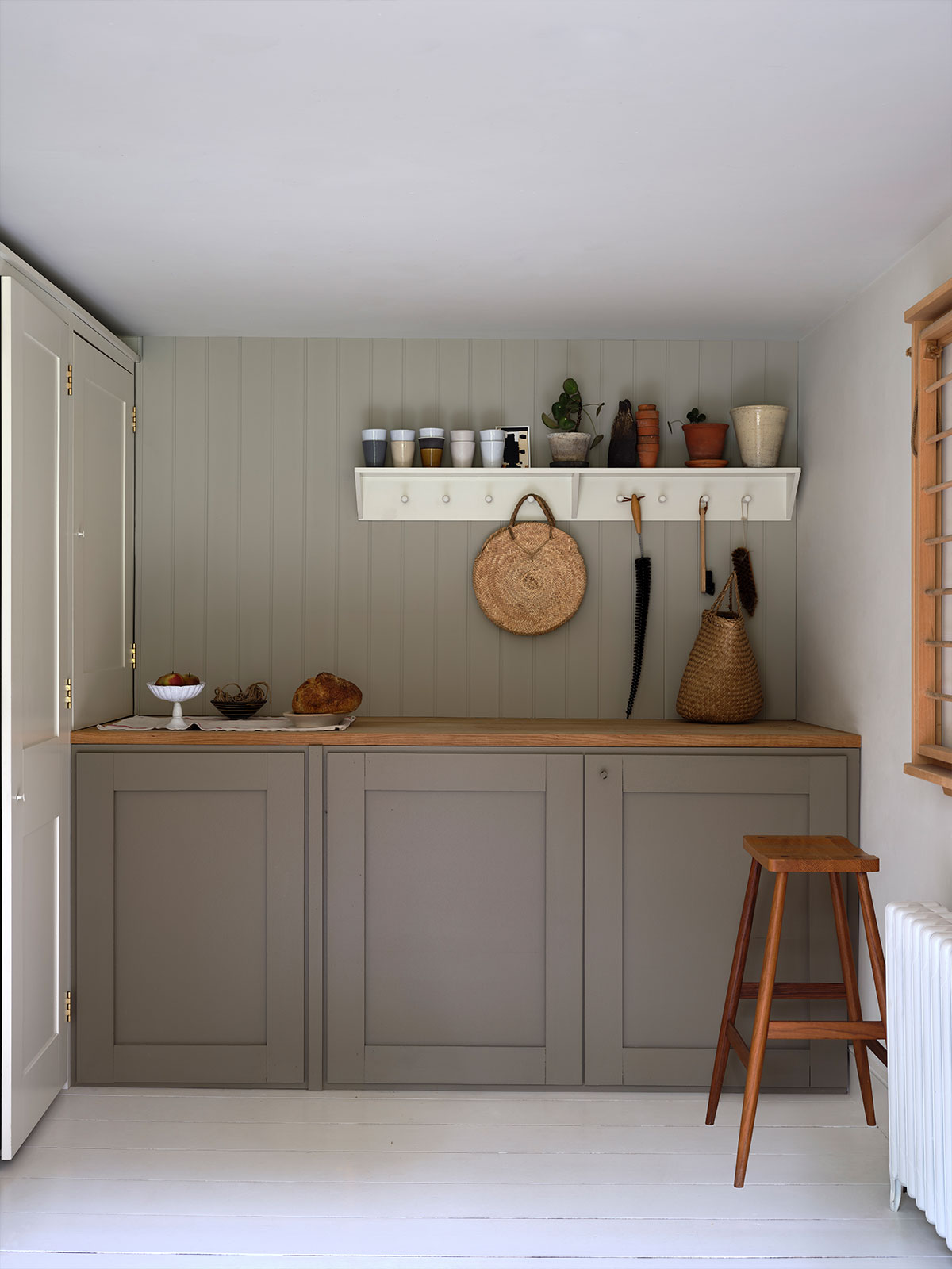 If you were seated on the wooden stool, you’d be surrounded on one side by the wooden worksurface above the cabinets. Perhaps the loaf of bread and peaches suggest it’s time to start preparing a simple, rustic lunch. More ingredients could be hidden behind the painted wooden doors, or in the large basket that’s just been put on the bench.