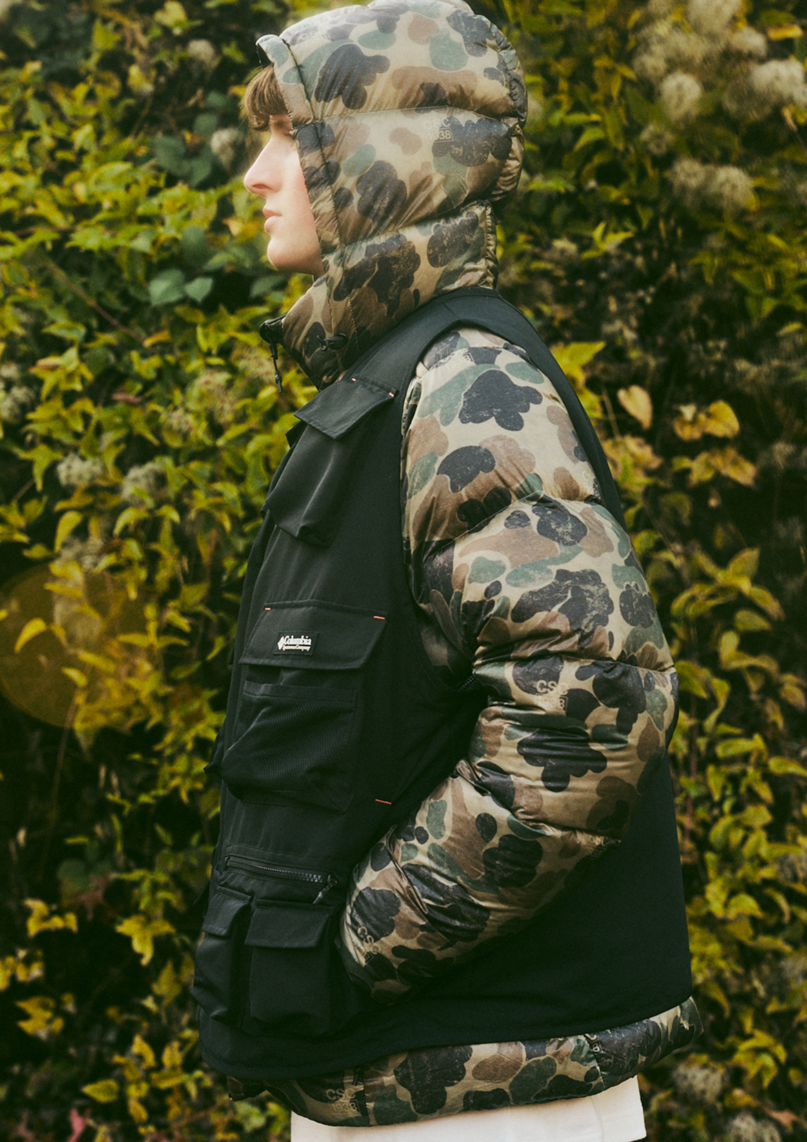 Side profile of man in CSC1938 Dually Mountain Printed Outerchange Jacket in camo and black, standing in front of greenery.