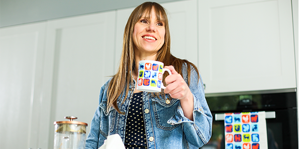Image of a woman smiling in her kitchen whilst holding a branded RSPCA mug. There is also a branded RSPCA tea towel in the background