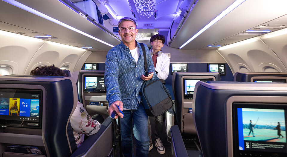 Two smiling men in the aisle of an aircraft with their carry-on luggage.