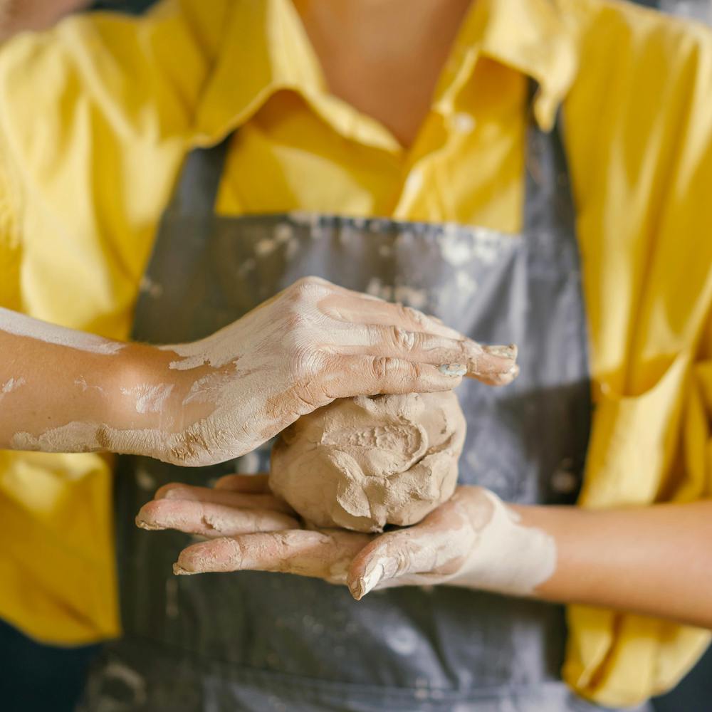 A person wearing a yellow shirt and blue apron holds a ball of clay between their hands.
