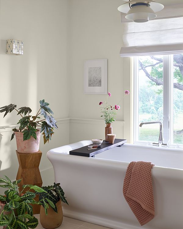 A white painted bathroom with a large rectangular tub, potted plants, and an open window.