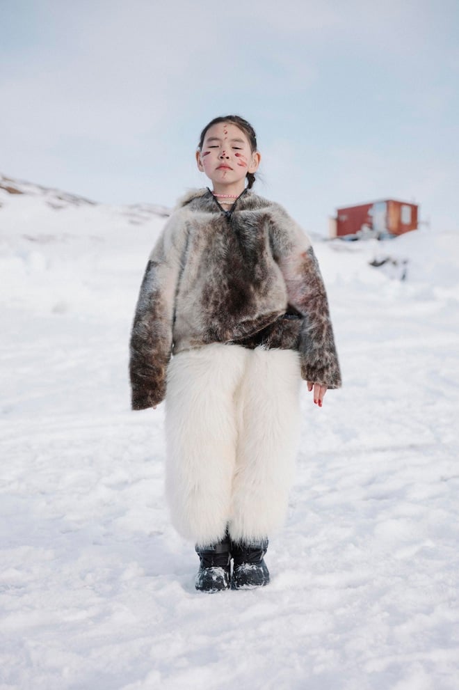 A colour photograph of a young Inuit girl standing on the sea ice of northern Greenland wearing traditional clothing made of seal fur and thick white fur pants. Her eyes are closed and her face is smeared with red seal blood, part of a local ritual performed after a seal hunt. She wears a beaded necklace and stands with a calm, composed posture. In the background, a red wooden cabin is visible on the island of Ikeresak.