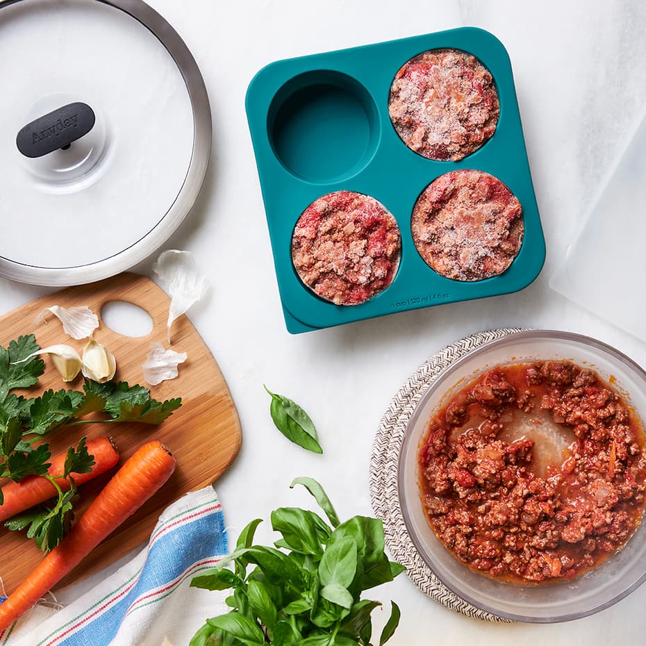 Bolognese sauce is stored in a kale-colored Anytime Freezer Tray, with the lay-flat silicone lid set aside. The tray sits next to an Anyday glass dish filled with more bolognese, surrounded by fresh ingredients like carrots, garlic, basil, and parsley on a white surface.