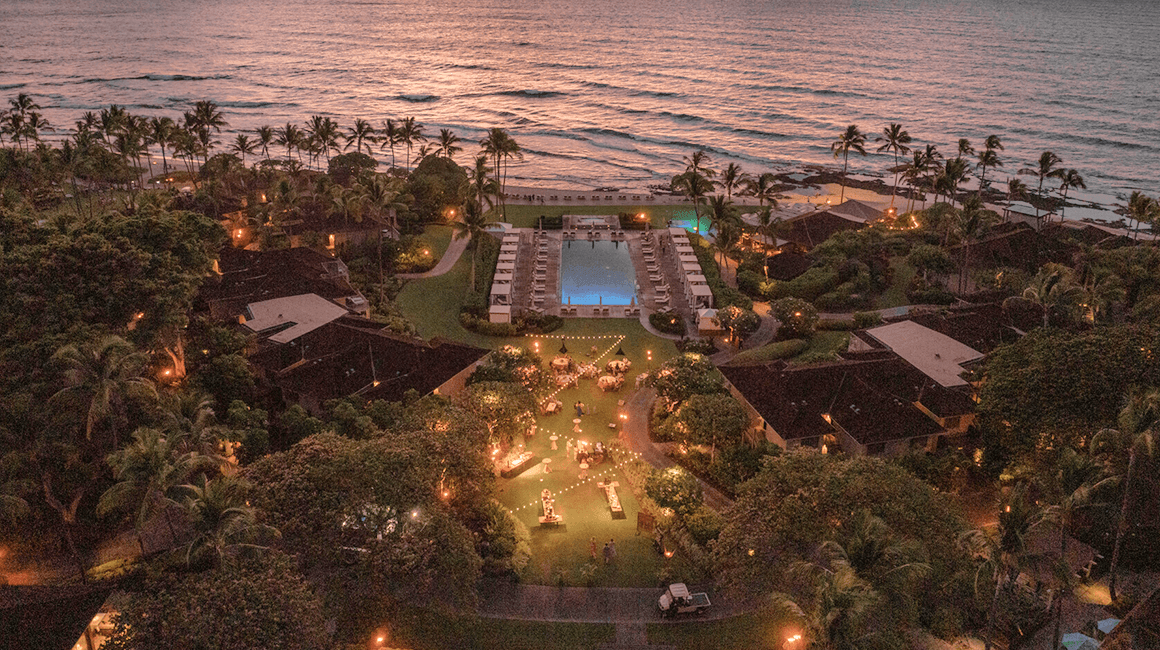 'Aerial view of a resort lit with decorative lights, with a pool and the ocean in the background.