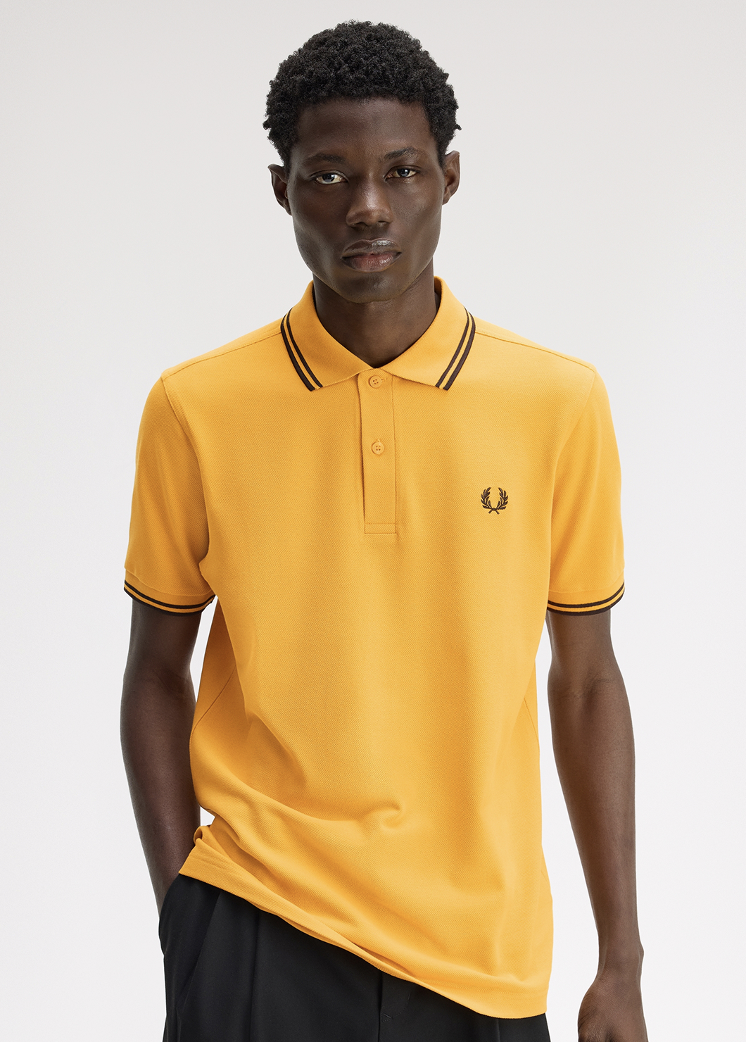 Black male model with short coily hair looks straight into the camera and wears a Fred Perry M3600 Twin Tipped Shirt in Golden / Rich Brown / Rich Brown, while he tucks one hand into his pocket