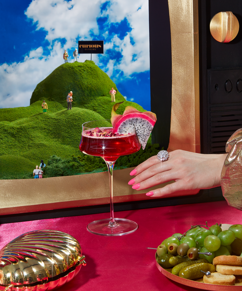 A coupe glass full of a pink drink sits in front of a gold TV displaying rolling green hills, miniature people, and a sign that says 