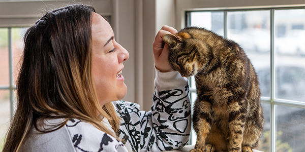 A woman wearing a white and black jumper is smiling at the tabby cat she is petting, who is sat on a cat tree by a window inside her living room