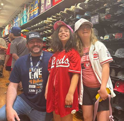 A man wearing a Lids Foundation hat and The Arc-branded shirt kneels alongside two young girls wearing Cincinnati Reds jerseys and caps, all smiling inside a Lids store surrounded by hats and sports jerseys.