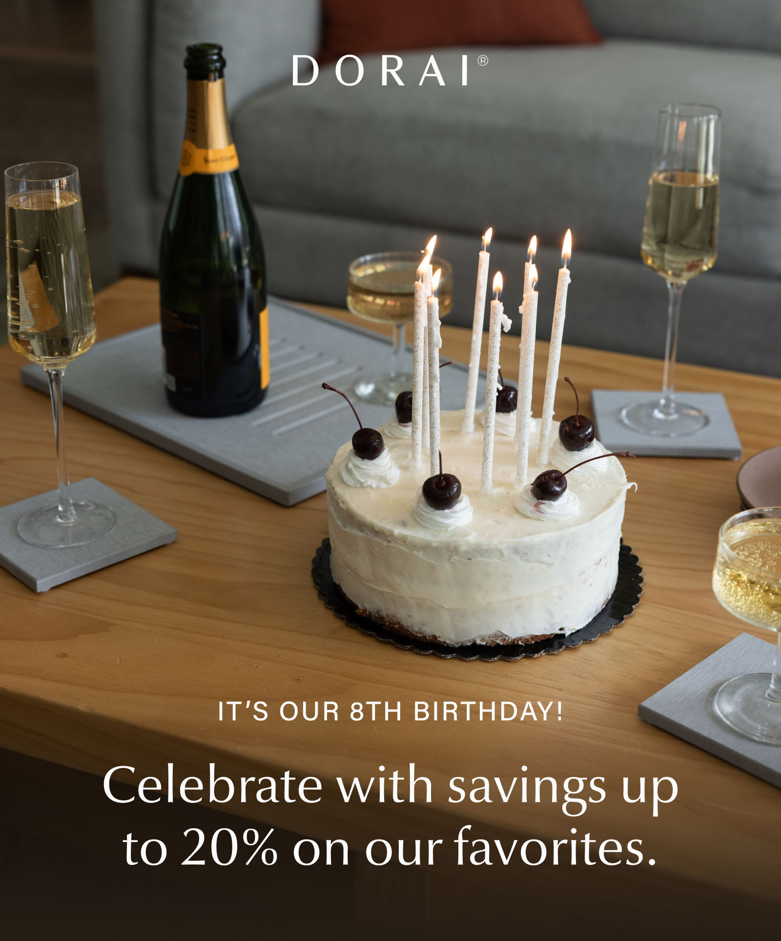 Birthday cake with lit candles on a coffee table surrounded by Dorai stone coasters and a stone serving tray, alongside glasses of sparkling wine and a champagne bottle. Text reads: "It's Our 8th Birthday! Celebrate with savings up to 20% on our favorites."