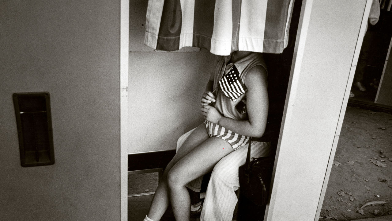 View, from the neck down of a young girl with American flag sitting on another person's lap as they pose behind the curtain of a photo booth