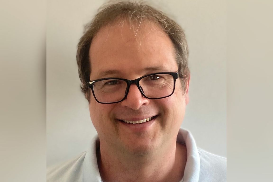 Headshot of Tommaso Treu, a man with brown hair, wearing black rectangular-framed glasses and a light blue collared shirt, smiling warmly against a neutral gray background