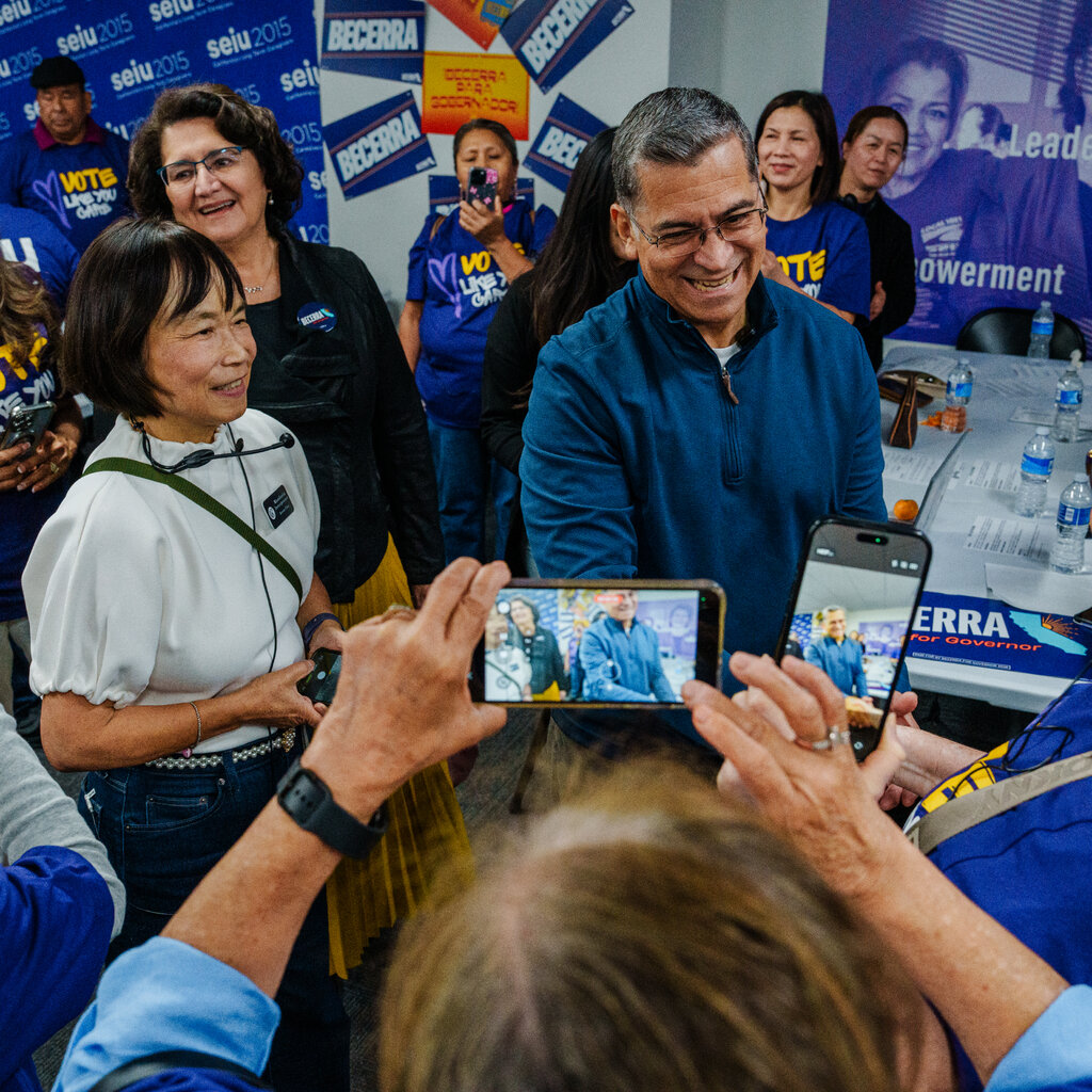 Xavier Becerra in a blue fleece, surrounded by a smiling crowd, many of them dressed in blue taking photos.