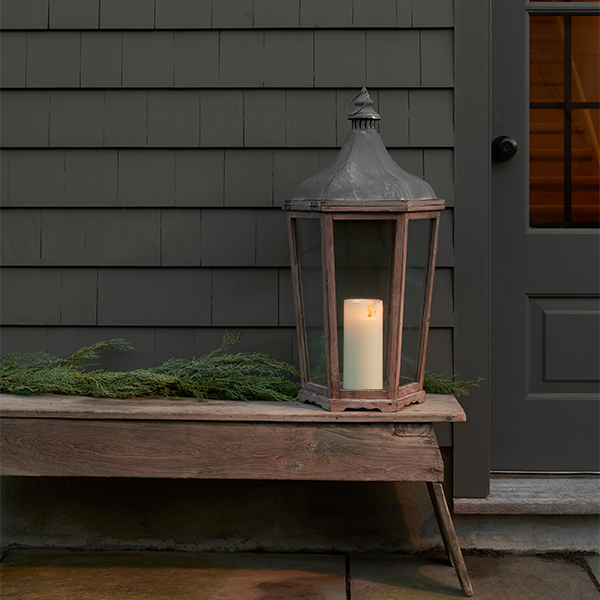 A home exterior with black siding and decorative wood bench topped with greenery and a lantern with a candle inside.