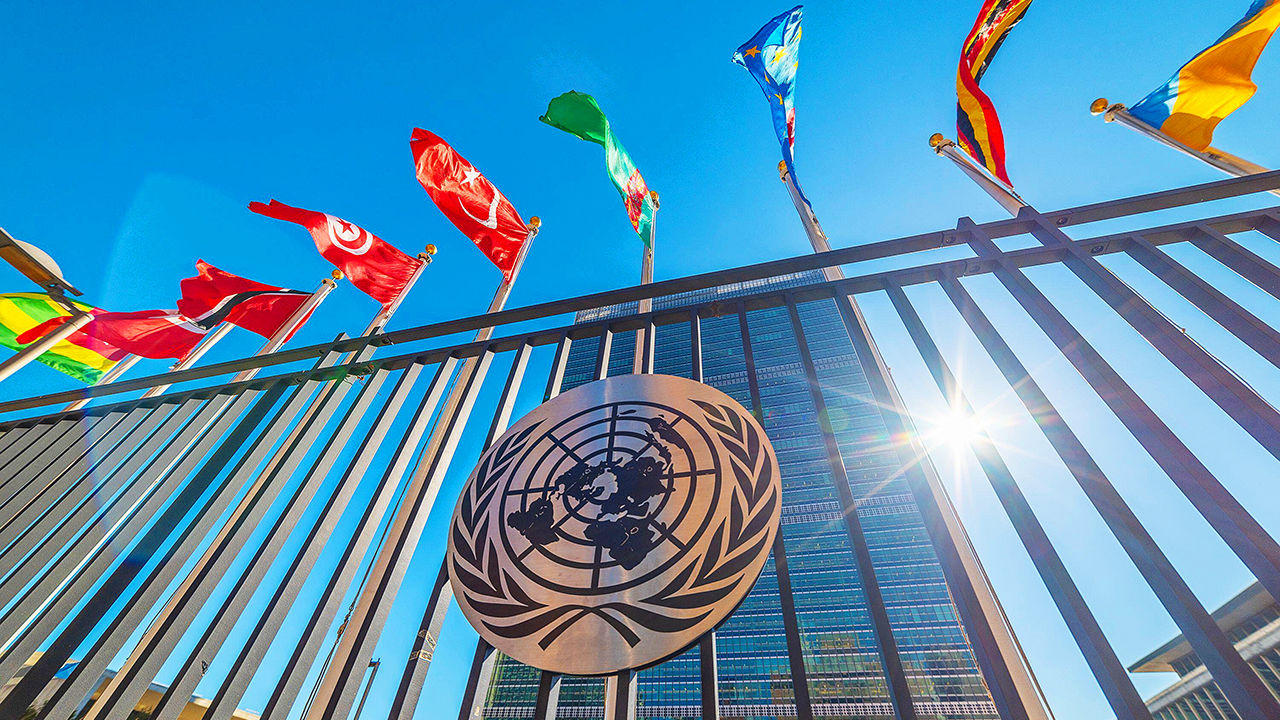 The headquarters of the United Nations building in New York - flags and UN logo in the foreground and blue sky in the background.