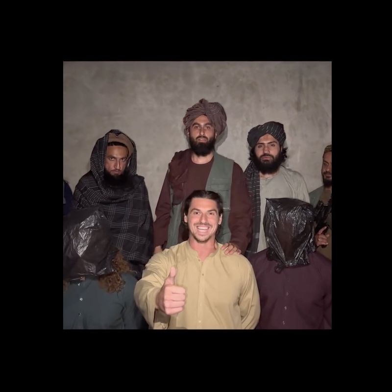 Screenshot image of Yosaf Aryubi and other men in turbans and tunics standing over kneeling men, center man is smiling with a thumbs up at the camera.