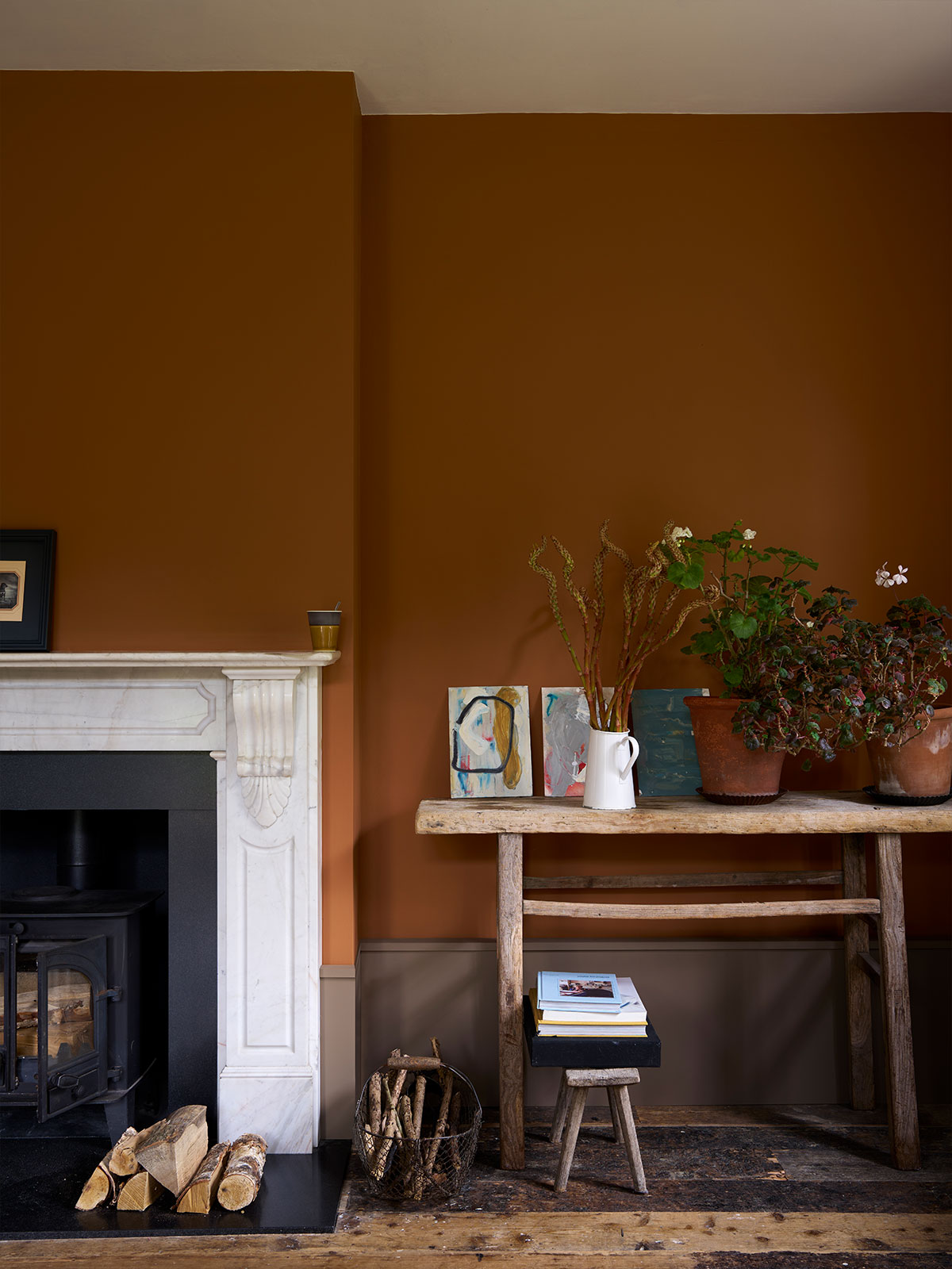 A warm living room corner with rich terracotta walls, a white marble fireplace and rustic wooden furniture. Potted plants, artwork and stacked books add character, creating a cosy and grounded atmosphere.