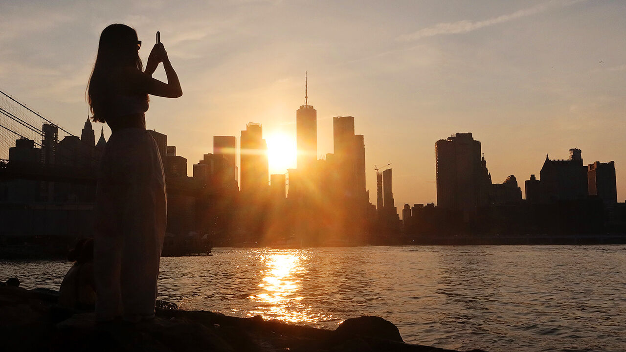 A woman photographs the sun setting behind the skyline of lower Manhattan and One World Trade Center from Brooklyn Bridge Park next to the East River in New York City, USA.