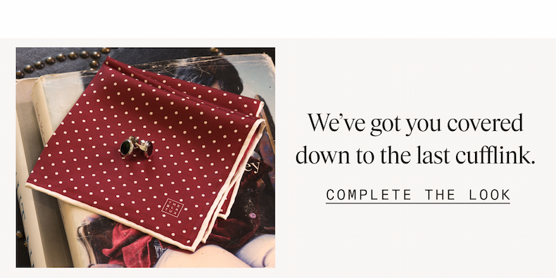 A burgundy polka dot pocket square with white trim, stamped with The Black Tux logo, resting atop stacked books alongside a gold and black cufflink. Text reads: "We've got you covered down to the last cufflink. COMPLETE THE LOOK"