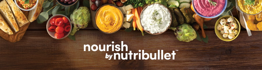 A top-down view of a wooden table with various healthy foods, including hummus dips, artichokes, olives, carrots, and sliced bread. The "nourish by nutribullet" logo is featured in white text.
