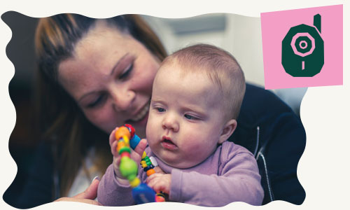 A baby being held by their smiling mum while playing with a toy
