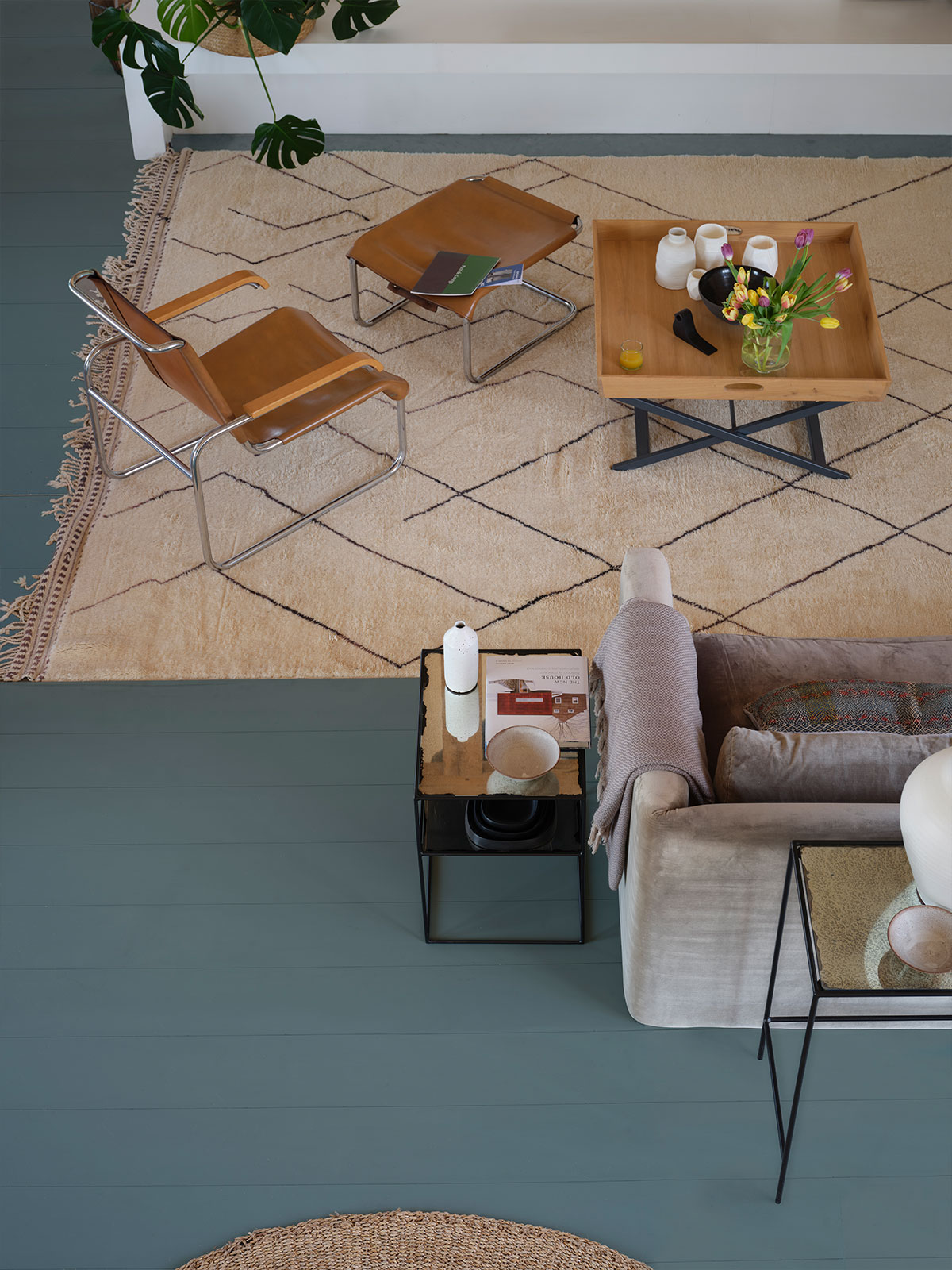 As we look down from an upper floor, we see a very bold statement flooring scheme. Petrol blue wooden flooring contrasts with a large, neutral-toned, fringed rug. A choice of seating with a tan leather chair with its own footstool or a grey velvet sofa make it a space you know has been designed for relaxing.