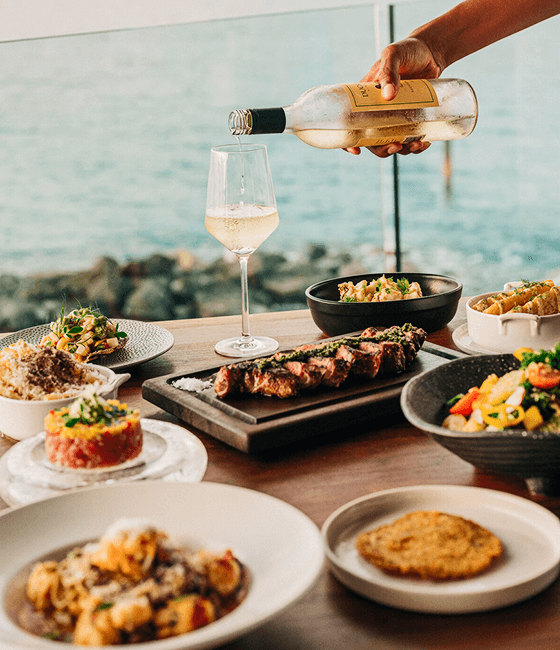 A table with gourmet dishes and a glass of white wine being poured by the sea.