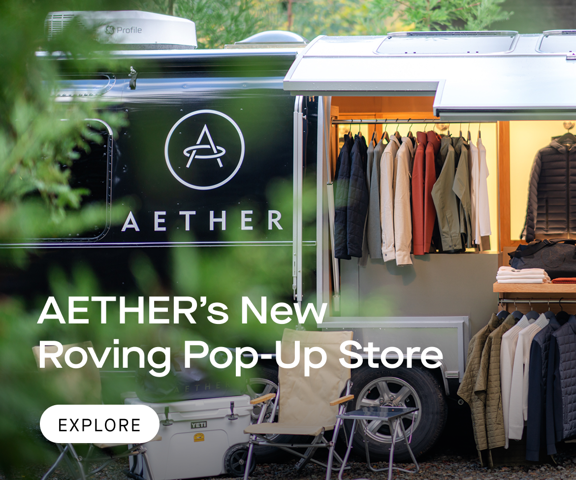 Image of the AETHER airstream with clothing display and two camping chairs setup in a forest environment.