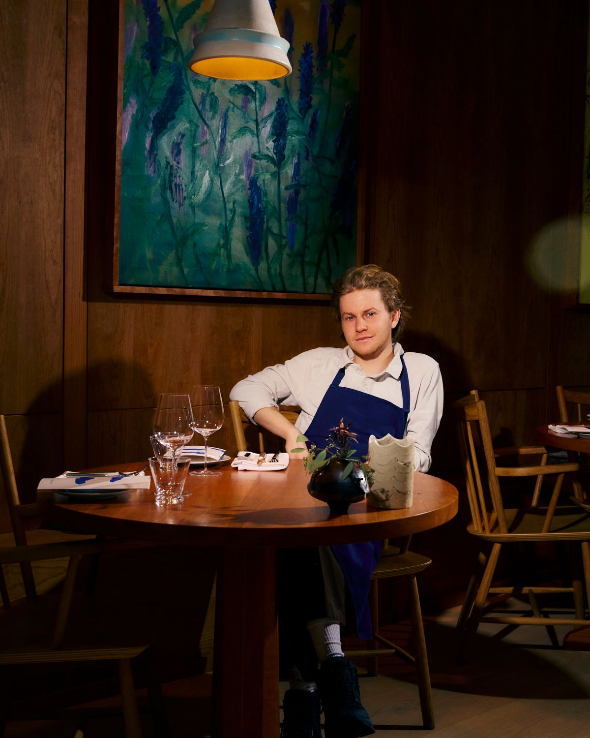 Flynn McGarry sitting by wooden dining table in his restaurant, with a painting behind him.