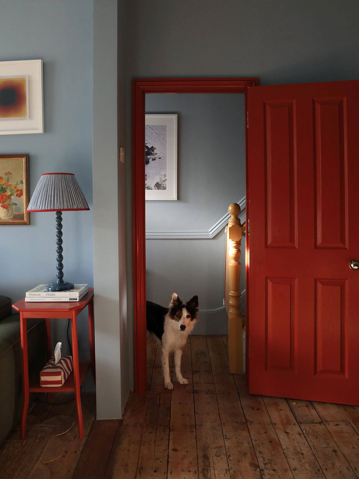 The dog looks inquisitively at you, almost as though it wants to know if you’re going to play. There’s definitely a cheerful playfulness to the surroundings, with the bright red of the door picked up on the trim of the lamp and the painted wooden table. By contrast, the soft blue of the walls is reminiscent of an early morning clear sky. With its bright paintings on the wall, it is not a room you could feel sad in.