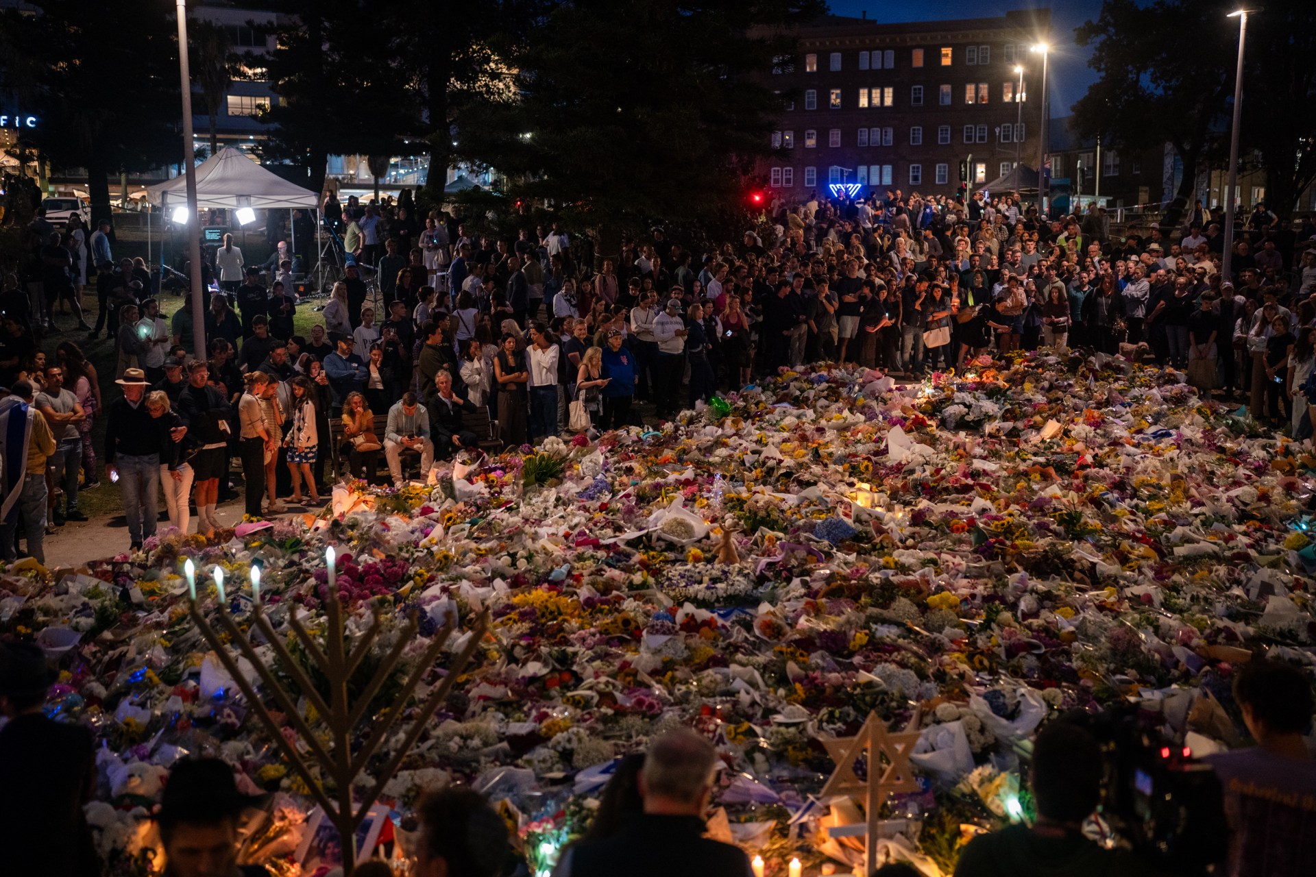 Mourners hold candles and gather around a display of flowers. 