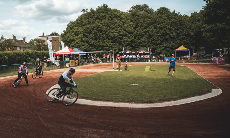 Three cycle speedway riders approach a corner at an event, with a judge in the centre of the track.
