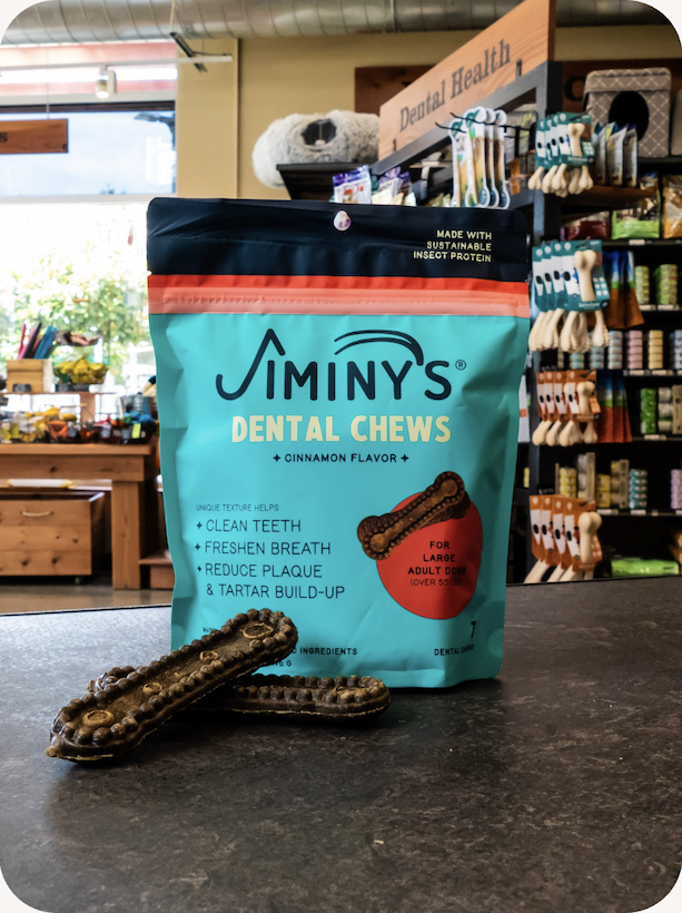 A photo of a bag of Jiminy's Dog Dental Chews on the front counter inside a Mud Bay location with two of the chews outside the bag. 