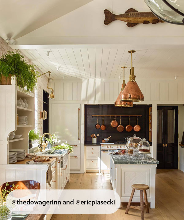 A serene country kitchen featuring cream cabinetry, copper pendants, marble counters, and hanging pans. Sunlight and warm timber floors infuse the space with an inviting sense of comfort and quiet rustic charm.