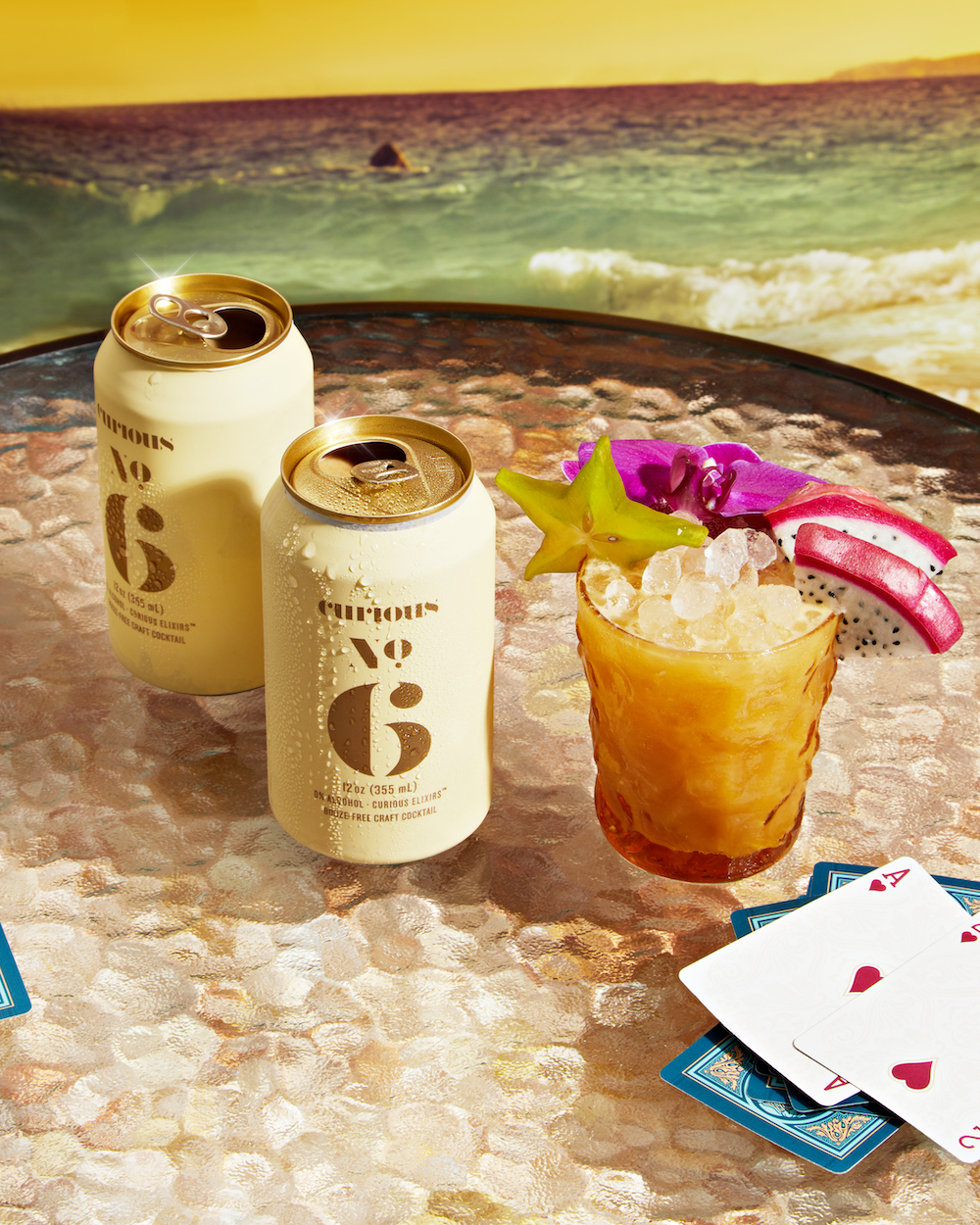 Two cans and a glass of Curious No. 6. The glass is garnished with tropical fruits. These all sit on a round glass table beside a stack of playing cards, with the ocean in the background.
