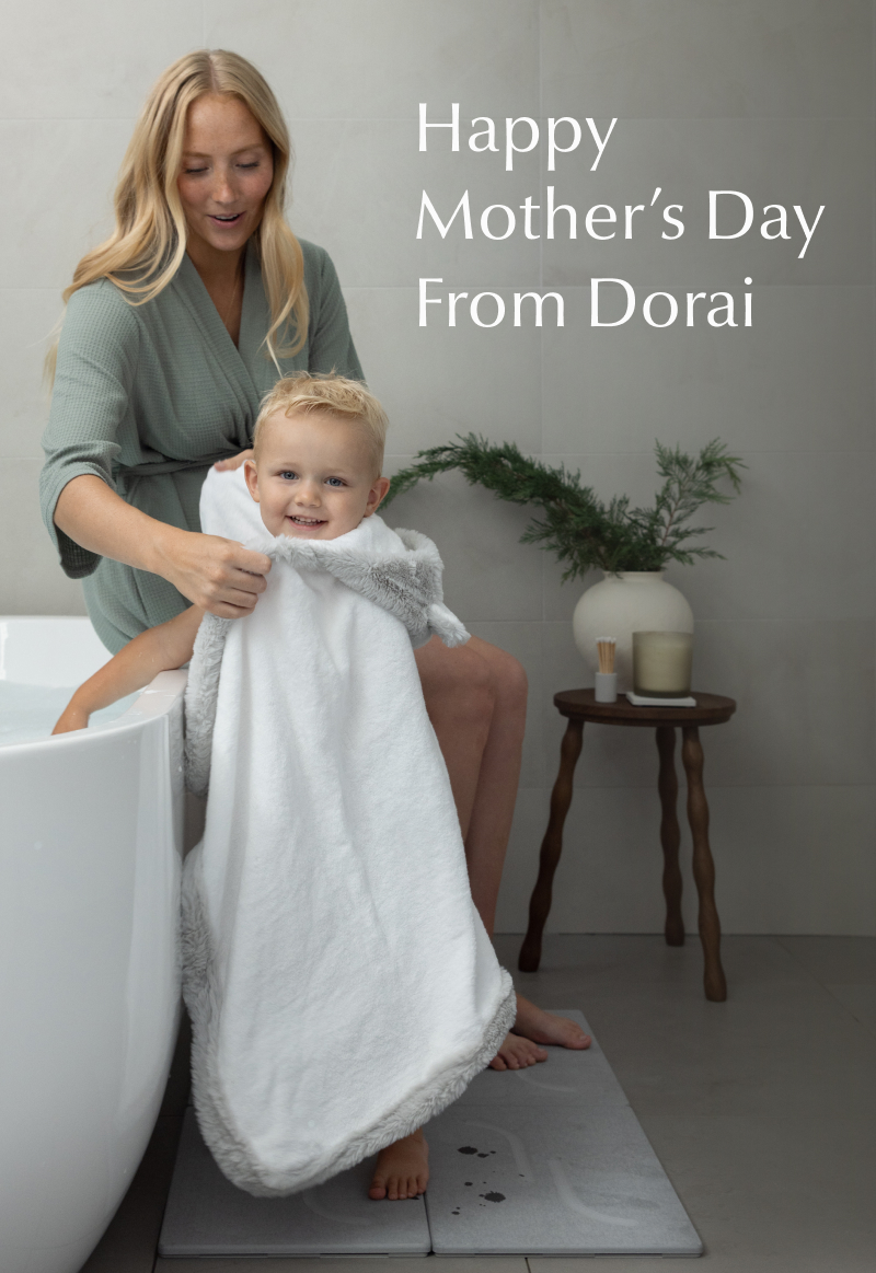 Mother in a soft green robe wraps a smiling toddler in a white towel beside a bathtub, standing on a gray stone bath mat. Text reads: “Happy Mother’s Day From Dorai.”