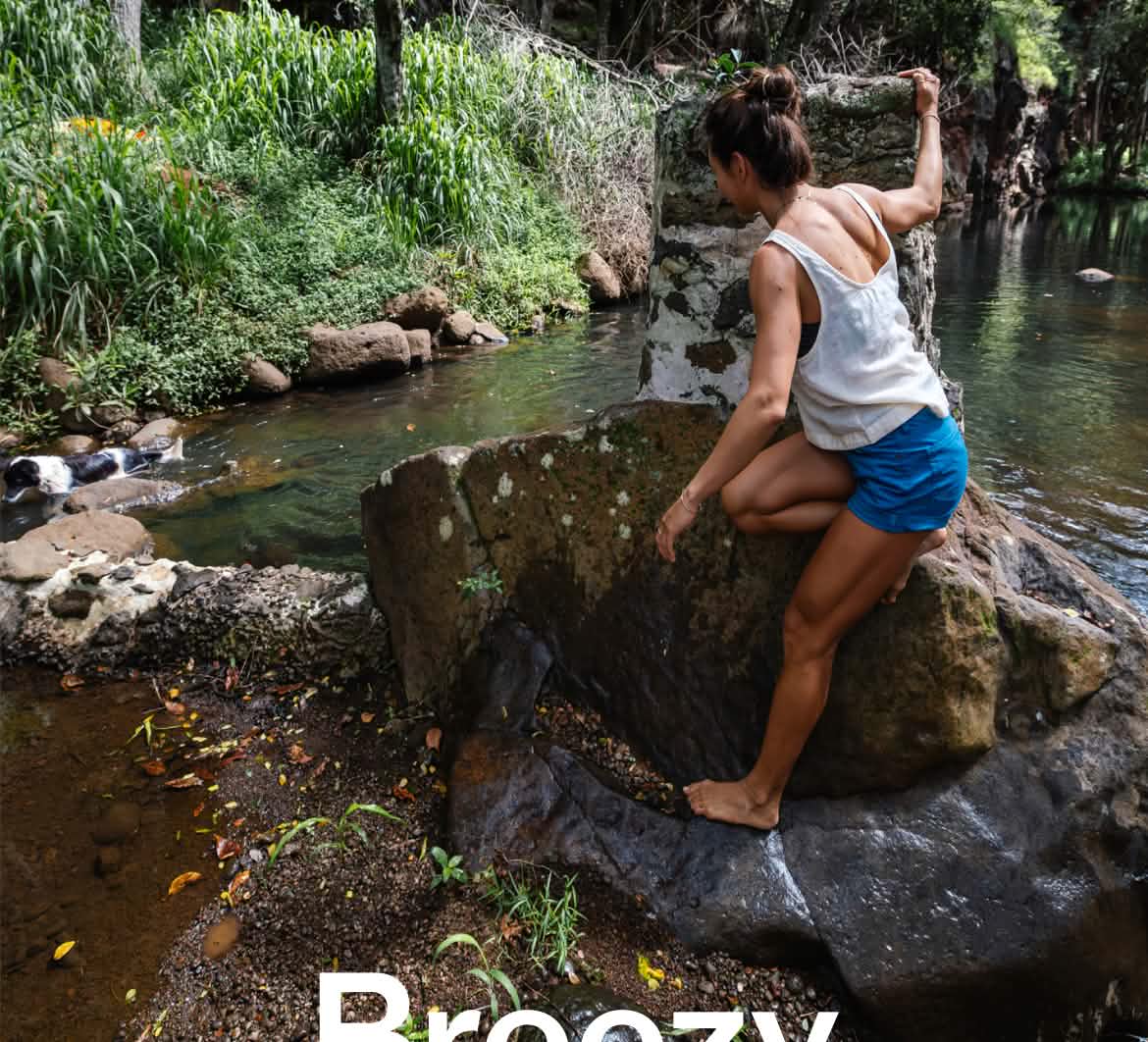 A person in shorts and a tank top leans against a rock by a watering hole surrounded by jungle.