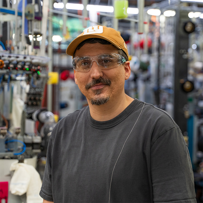 Kris M smiles in front of a row of knitting machines